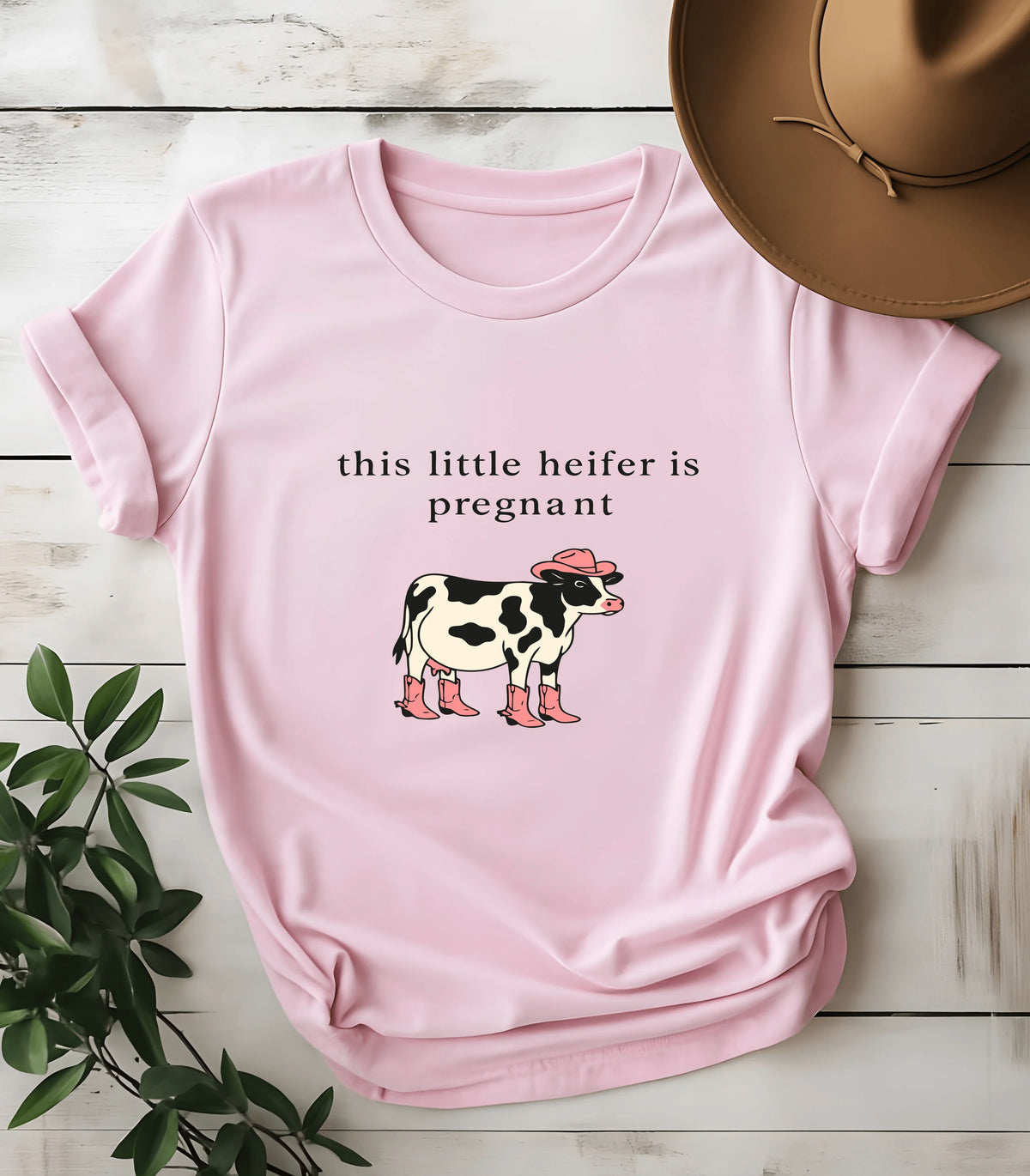 Pink graphic tee with 'this little heifer is pregnant' text and cow in cowboy hat and boots