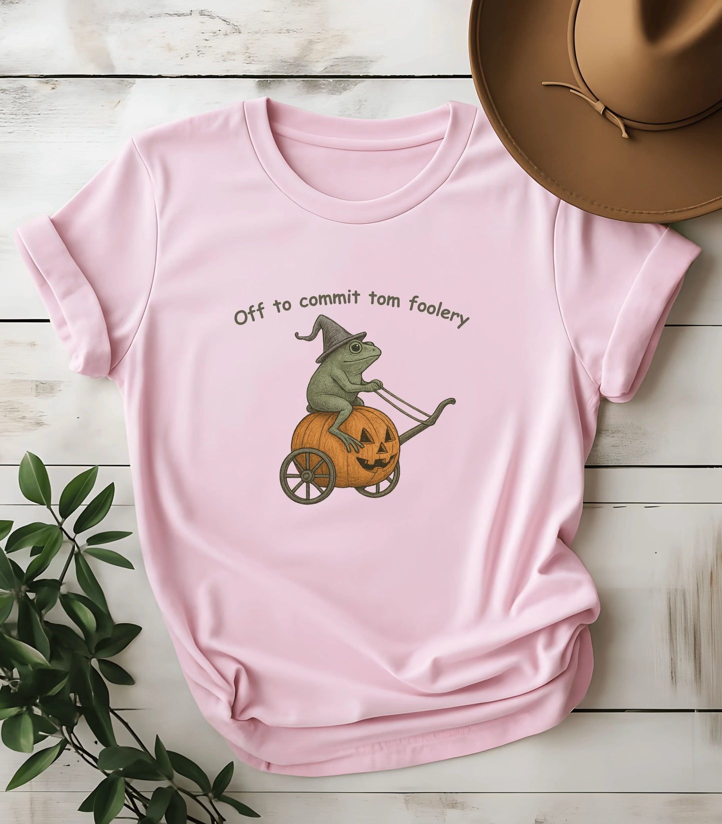 Pink t-shirt with frog in witch hat riding pumpkin cart, text 'Off to commit tom foolery'
