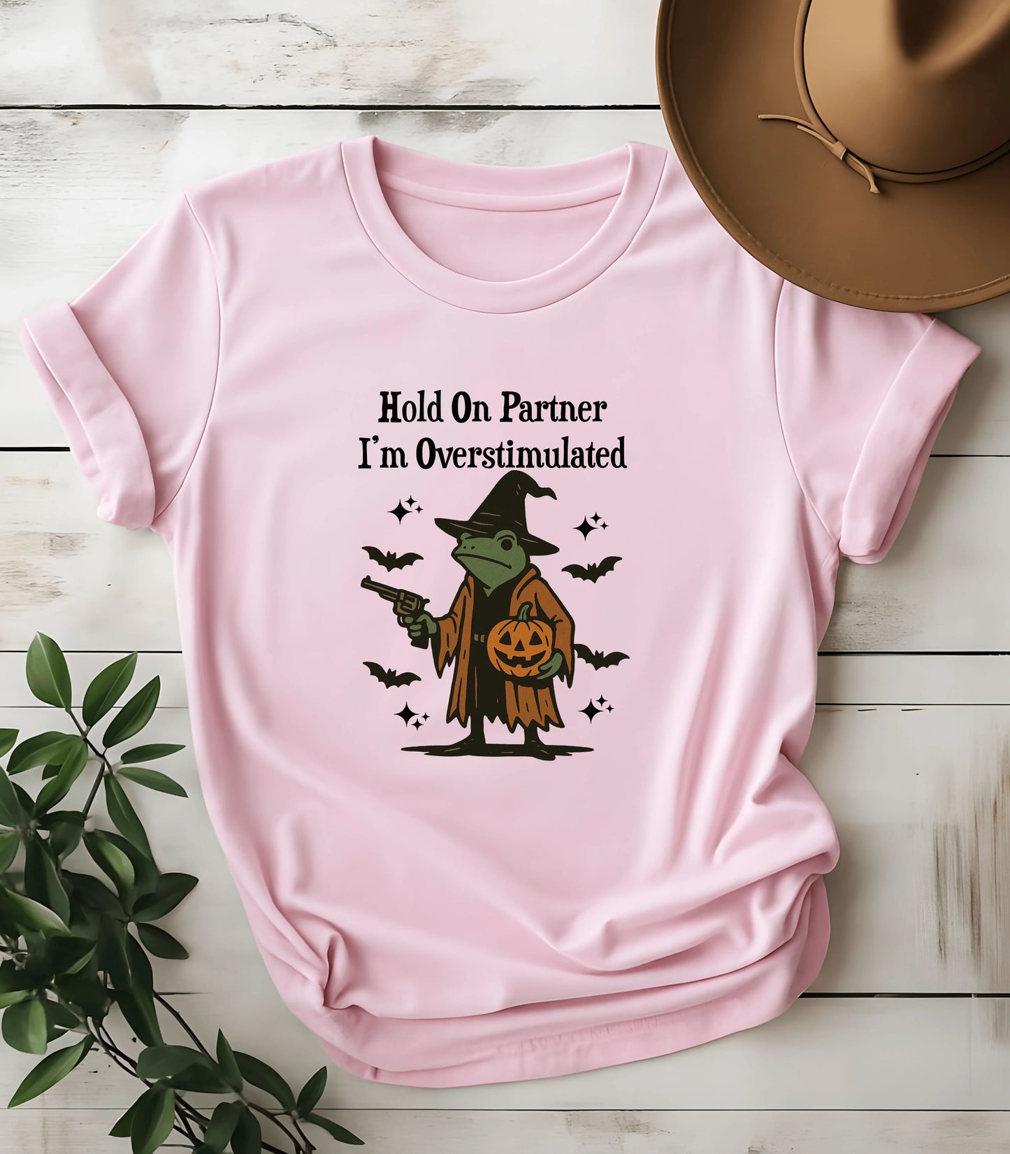 Pink graphic tee with witch frog holding pumpkin and gun, text 'Hold On Partner I'm Overstimulated'